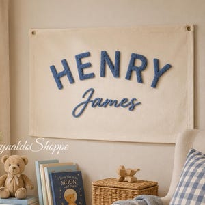 May include: A rectangular cream-colored fabric wall hanging featuring the name HENRY JAMES in raised, textured blue lettering. The word HENRY is in a bold, block-style font, while the name James is in a cursive, script-style font.