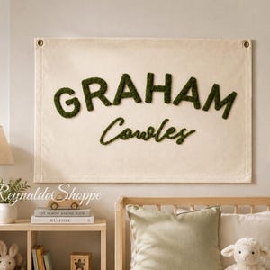 May include: A rectangular off-white canvas wall hanging featuring the name GRAHAM Cowles in raised, dark green textured lettering. The fabric banner includes metal grommets in the top corners for mounting and displays a custom name design.