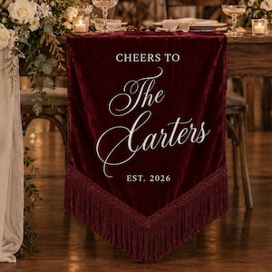 May include: A deep burgundy velvet banner featuring white text that reads CHEERS TO The Carters EST. 2026. The banner has a pointed bottom edge finished with matching burgundy fringe trim and is displayed hanging from the edge of a wooden table.