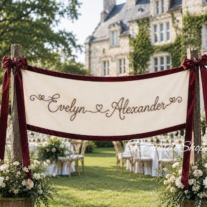 May include: A cream-coloured fabric banner featuring the embroidered names Evelyn and Alexander in dark brown script, separated by a heart. The banner is trimmed in deep red velvet, finished with matching bows on two wooden posts.