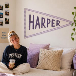 Könnte beinhalten: Ein dreieckiger Wandwimpel mit dem Namen HARPER in großen violetten Buchstaben und einem violetten Karomuster am Rand. Der Wimpel hängt an einer schlichten Wand über einem Bett mit verschiedenen Kissen, darunter ein strukturiertes beiges Kissen.