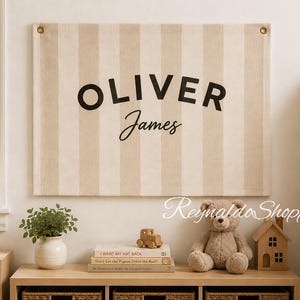 May include: A rectangular wall hanging featuring vertical beige and cream stripes with the name OLIVER JAMES printed in black lettering. The fabric banner includes metal grommets in the top corners for mounting, displaying a clean, neutral aesthetic.