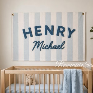 May include: A rectangular wall hanging featuring vertical light blue and white stripes with the name HENRY in dark blue block letters and Michael in dark blue script lettering. The fabric banner is mounted with metal grommets in the top corners.