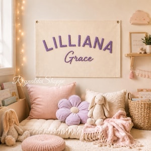 May include: A rectangular cream-colored wall hanging featuring the name LILLIANA in purple block letters and the name Grace in purple script lettering. The fabric banner is mounted with metal grommets at the top corners.