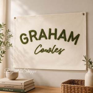May include: A rectangular off-white canvas wall hanging featuring the name GRAHAM in large, dark green, textured, tufted block letters, with the name Cowles in smaller, dark green, cursive, tufted script lettering below the first name.