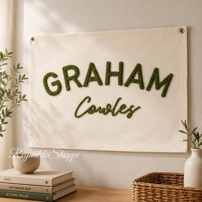 May include: A rectangular off-white canvas wall hanging featuring the name GRAHAM in large, dark green, textured, tufted block letters, with the name Cowles in smaller, dark green, cursive, tufted script lettering below the first name.