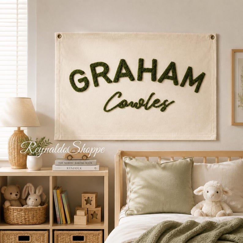 May include: A rectangular off-white canvas wall hanging featuring the name GRAHAM Cowles in raised, dark green textured lettering. The fabric banner includes metal grommets in the top corners for mounting and displays a custom name design.
