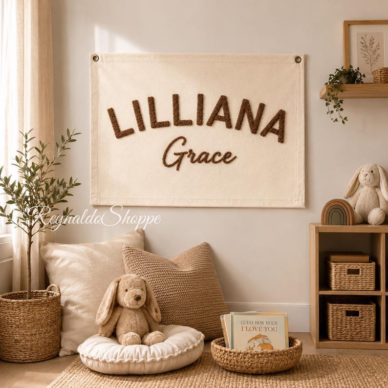 May include: A rectangular cream-colored fabric wall hanging featuring the name LILLIANA Grace in raised, dark brown textured lettering. The banner includes metal grommets in the top corners for mounting and is displayed on a plain wall.