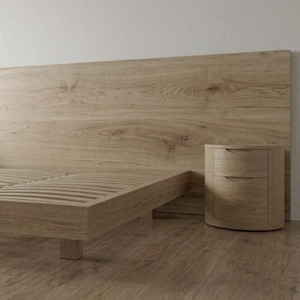 Puede incluir: Un marco de cama de madera de roble claro con un cabecero rectangular ancho que se extiende horizontalmente detrás de dos mesitas de noche cilíndricas a juego. La estructura de la cama incluye una base de listones y patas cortas.