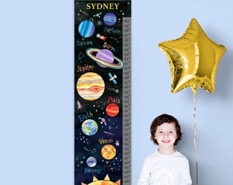 May include: A colorful growth chart featuring a black background with illustrations of planets and the sun. The chart is labeled with the name "SYDNEY" and includes height measurements in inches. A young boy is sitting on a white bench in front of the chart.