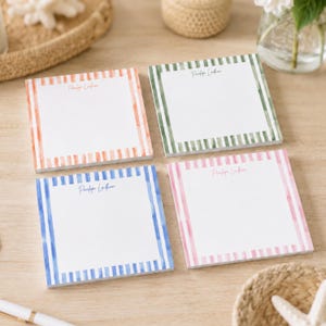 Könnte beinhalten: Vier quadratische Haftnotizblöcke mit gestreiften Rändern im Aquarellstil in Orange, Grün, Blau und Pink. Jeder Block trägt den Namen Penelope Lindham, der in einer Schreibschrift oben mittig auf der weißen Schreibfläche gedruckt ist.