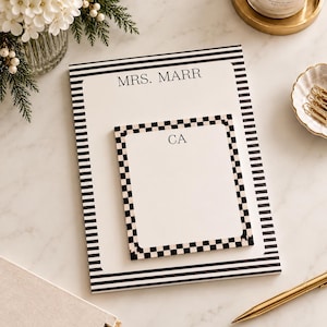May include: A rectangular notepad with black and white horizontal stripes at the top and bottom, featuring the text MRS. MARR. A smaller square notepad with a black and white checkered border and the initials CA sits on top of the larger pad.