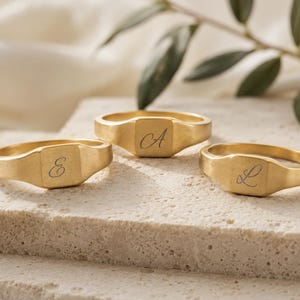 May include: Three gold-toned signet rings with a matte finish, each featuring a single engraved cursive letter on the flat square face. The letters E, A, and L are clearly visible on the respective rings, which are arranged in a cluster.