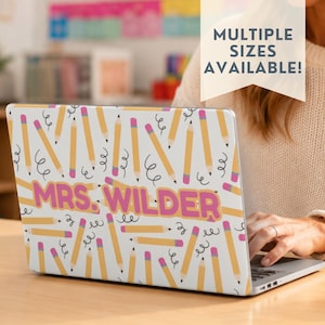 Könnte beinhalten: Eine Laptop-Hülle mit weißem Hintergrund und einem sich wiederholenden Muster aus gelben Bleistiften mit rosa Radiergummis und schwarzen Kringeln. Der Text MRS. WILDER ist in großen, fetten rosa Buchstaben mittig aufgedruckt.