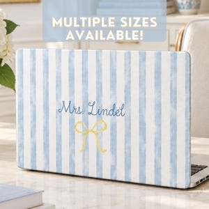 Peut inclure: Un skin pour ordinateur portable avec un motif à rayures aquarelle bleu clair et blanc. Le texte "Mrs. Lindel" est imprimé en écriture manuscrite bleu foncé au-dessus d'un nœud jaune dessiné à la main. Une bannière bleue affiche "MULTIPLE SIZES AVAILABLE!"