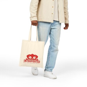 May include: A cream-colored canvas tote bag featuring a red graphic of a dog peeking over the text "INTROVERTED but will discuss books" printed in red. The bag has two long shoulder straps and a rectangular shape made of woven fabric.