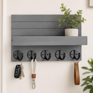 Puede incluir: Un organizador de pared de madera gris con un estante horizontal y cinco ganchos de metal negro. El estante sostiene una pequeña planta verde en maceta. Dos ganchos tienen llaveros, uno con una llave de coche negra y otro con una correa de cuero marrón.