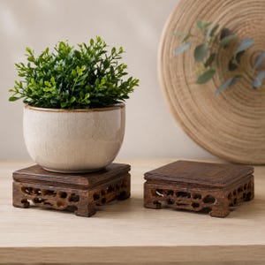 May include: A small, round, off-white ceramic pot with a subtle crackle glaze finish containing a lush green artificial plant, resting on a square, dark brown wooden display stand with intricate, carved openwork detailing on the sides.