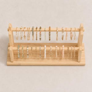 May include: A two-tiered light wood jewelry display stand holding a collection of delicate bracelets and rings. The stand features two horizontal bars supported by side frames, showcasing various beaded, metallic, and gemstone jewelry pieces.