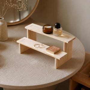 Può includere: Un espositore a due livelli in legno chiaro dal design rettangolare con supporti verticali. Il ripiano superiore ospita un barattolo di candela in vetro marrone e un flacone di profumo in vetro trasparente, mentre quello inferiore mostra un orecchino a cerchio dorato e un cofanetto quadrato.