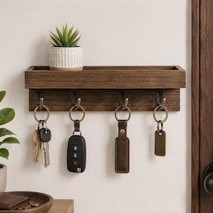 May include: A dark brown wooden wall-mounted shelf with four metal hooks. Each hook holds a key ring with various keys, a car key fob, a brown leather keychain, and a blank metal tag. A small potted succulent sits on the top shelf.