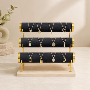 May include: A three-tier jewelry display stand featuring black velvet-covered cylindrical bars supported by gold-toned metal rods on a light wood base, holding twelve gold necklaces with various pendants including a heart, lock, and moon.