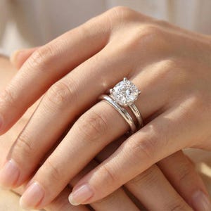 Puede incluir: Un conjunto de anillos de compromiso de tono plateado con una piedra central grande, redonda y de corte brillante en un engaste de cuatro puntas, junto a una alianza de boda de tono plateado lisa, pulida y sencilla en el mismo dedo.