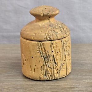 May include: A cylindrical wooden container with a matching lid, featuring a light tan color and a natural, textured surface with dark, irregular grain patterns and small holes throughout the exterior, showcasing the raw wood grain and texture.