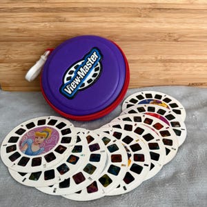 May include: A purple circular View-Master storage case with a red zipper and a white pull tab, accompanied by a collection of white circular image reels. One reel features a graphic of the character Cinderella in a blue dress on a pink background.