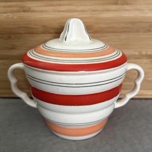 May include: A ceramic sugar bowl with a lid, featuring a cream-colored base with horizontal stripes in red and peach. The bowl has two side handles and a finial on the lid, with a visible crackle glaze finish throughout the entire surface.