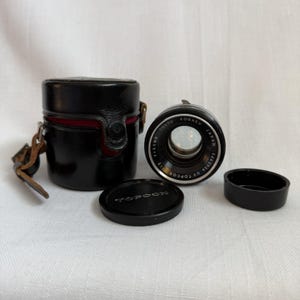 Puede incluir: Un objetivo de cámara negro con el texto Tokyo Kogaku Japan 53mm 1:2 UV Topcor 5465956, una tapa de objetivo negra con el texto Topcon, una segunda tapa negra y un estuche cilíndrico negro con interior rojo y una correa de cuero marrón.