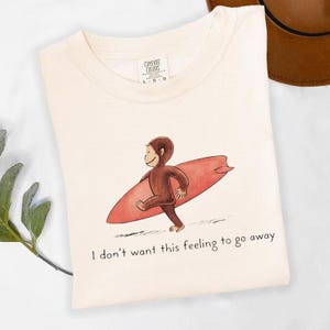 以下が含まれることがあります： クリーム色のTシャツ。赤いサーフボードを持った漫画の猿が描かれています。「I don't want this feeling to go away」の文字が下にプリントされています。Tシャツは折りたたまれており、Comfort Colorsのタグが付いています。