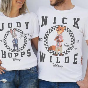 May include: White t-shirts featuring characters from Disney's Zootopia. One shirt displays Judy Hopps, the other Nick Wilde, each within a checkered circle. The shirts have the characters' names and the Disney logo.