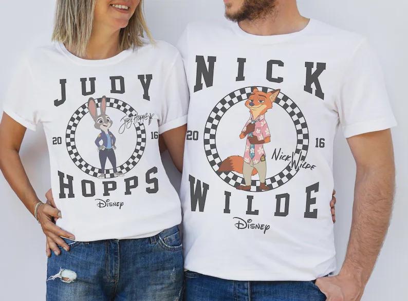 May include: White t-shirts featuring characters from Disney's Zootopia. One shirt displays Judy Hopps, the other Nick Wilde, each within a checkered circle. The shirts have the characters' names and the Disney logo.
