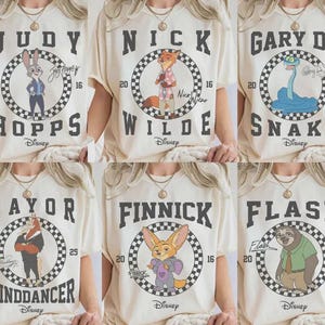 May include: Cream-colored t-shirts featuring characters from the Disney movie Zootopia. Each shirt displays a character's name in bold letters above a circular graphic with the Disney logo. Characters include Judy Hopps, Nick Wilde, and Gary Snake.