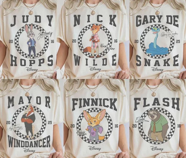 May include: Cream-colored t-shirts featuring characters from the Disney movie Zootopia. Each shirt displays a character's name in bold letters above a circular graphic with the Disney logo. Characters include Judy Hopps, Nick Wilde, and Gary Snake.