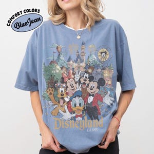 May include: Blue short-sleeved t-shirt with a Disneyland graphic featuring Mickey Mouse, Minnie Mouse, Donald Duck, and Goofy. The shirt has the text "Disneyland Est. 1955" in gold lettering. The shirt is labeled "Comfort Colors Blue Jean".