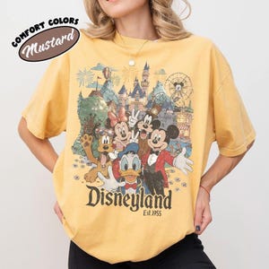 May include: Mustard-colored t-shirt featuring a Disneyland graphic with Mickey Mouse, Minnie Mouse, Donald Duck, Goofy, and other characters. The shirt has the text "Disneyland Est. 1955" in a vintage style. The words "Comfort Colors" and "Mustard" are also visible.