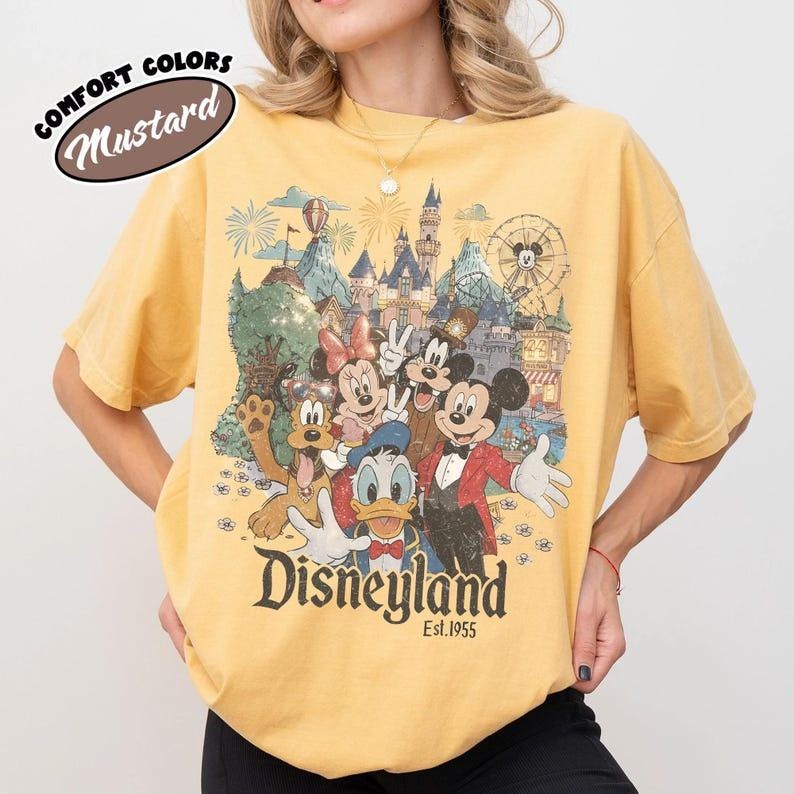May include: Mustard-colored t-shirt featuring a Disneyland graphic with Mickey Mouse, Minnie Mouse, Donald Duck, Goofy, and other characters. The shirt has the text "Disneyland Est. 1955" in a vintage style. The words "Comfort Colors" and "Mustard" are also visible.