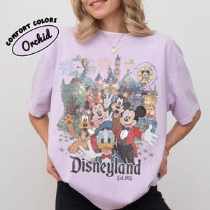 May include: A light purple t-shirt featuring a Disneyland graphic with Mickey Mouse, Minnie Mouse, Donald Duck, Goofy, and other characters. The shirt has the text "Disneyland Est. 1955" and "Comfort Colors Orchid" on the top left.