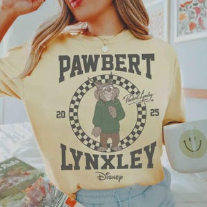 May include: Yellow t-shirt featuring a cartoon lynx character in a green sweater, with the text "PAWBERT LYNXLEY" in dark gray. The design includes a checkered circle and the Disney logo. The shirt has a relaxed fit.