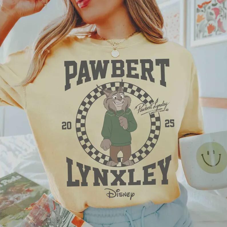 May include: Yellow t-shirt featuring a cartoon lynx character in a green sweater, with the text "PAWBERT LYNXLEY" in dark gray. The design includes a checkered circle and the Disney logo. The shirt has a relaxed fit.