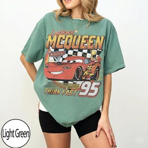 K&ouml;nnte beinhalten: Hellgr&uuml;nes T-Shirt mit einem Lightning McQueen-Motiv aus dem Film Cars. Der Aufdruck enth&auml;lt die Schriftz&uuml;ge "Lightning McQueen", "Cars", "Think Fast" und die Nummer "95". Das Design ist in Rot, Orange, Gelb und Schwarz gehalten.
