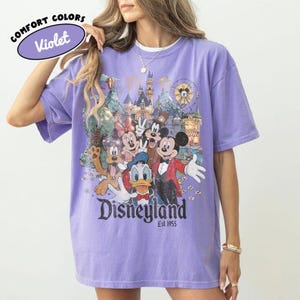 May include: Violet Comfort Colors t-shirt featuring a colorful Disneyland graphic with Mickey Mouse, Minnie Mouse, Donald Duck, and Goofy. The shirt has the text "Disneyland Est. 1955". The shirt is a light purple color.