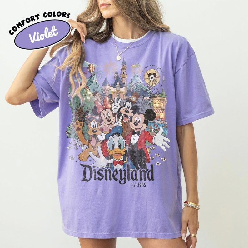 May include: Violet Comfort Colors t-shirt featuring a colorful Disneyland graphic with Mickey Mouse, Minnie Mouse, Donald Duck, and Goofy. The shirt has the text "Disneyland Est. 1955". The shirt is a light purple color.