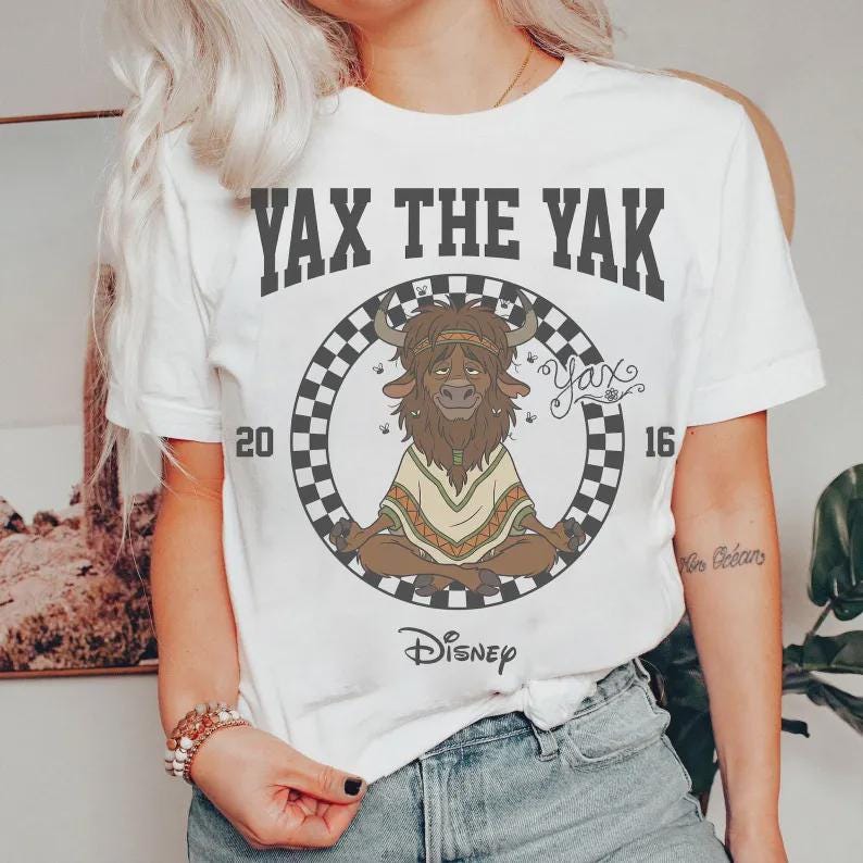 May include: White t-shirt featuring a cartoon illustration of Yax the Yak from Disney's Zootopia. The design includes the text "YAX THE YAK" above a circular graphic with the character in a meditative pose, and the Disney logo.