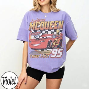 K&ouml;nnte beinhalten: Ein lila T-Shirt mit einer Grafik von Lightning McQueen aus dem Film Cars. Das Design enth&auml;lt den Text "Lightning McQueen", "Cars", "95" und "Think Fast".
