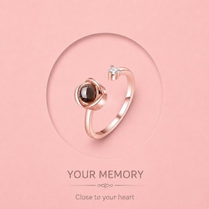 Puede incluir: Anillo de oro rosa con una piedra central marrón oscuro y una pequeña piedra transparente en el extremo abierto. El anillo está sobre un fondo rosa con el texto "YOUR MEMORY" y "Close to your heart".