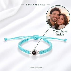Peut inclure: Un bracelet en cordon tressé bleu clair avec un médaillon métallique circulaire central doté d'une lentille de projection sombre. Le texte LUNAMYRIA est inscrit au-dessus, et un encart circulaire montre un couple souriant avec le texte Your photo inside.