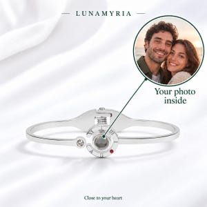 Op de afbeelding: Een zilverkleurige scharnierende armband met een centrale ronde bedel met een projectielens, een kleine heldere kristal en een rode accentsteen. De tekst LUNAMYRIA en Close to your heart zijn zichtbaar, samen met een ronde foto-inzet.